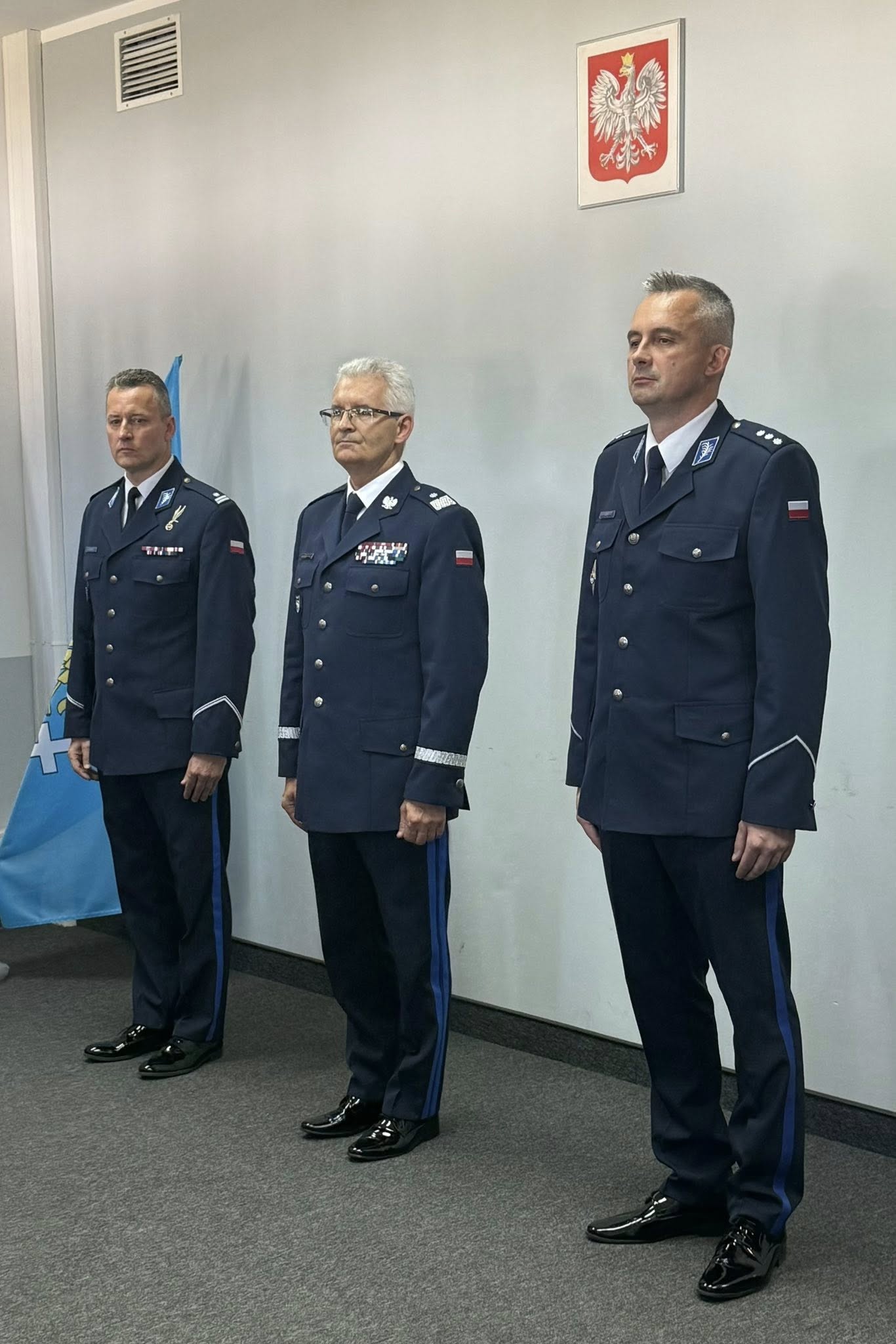 Trzej policjanci stojący w pomieszczeniu w mundurach galowych