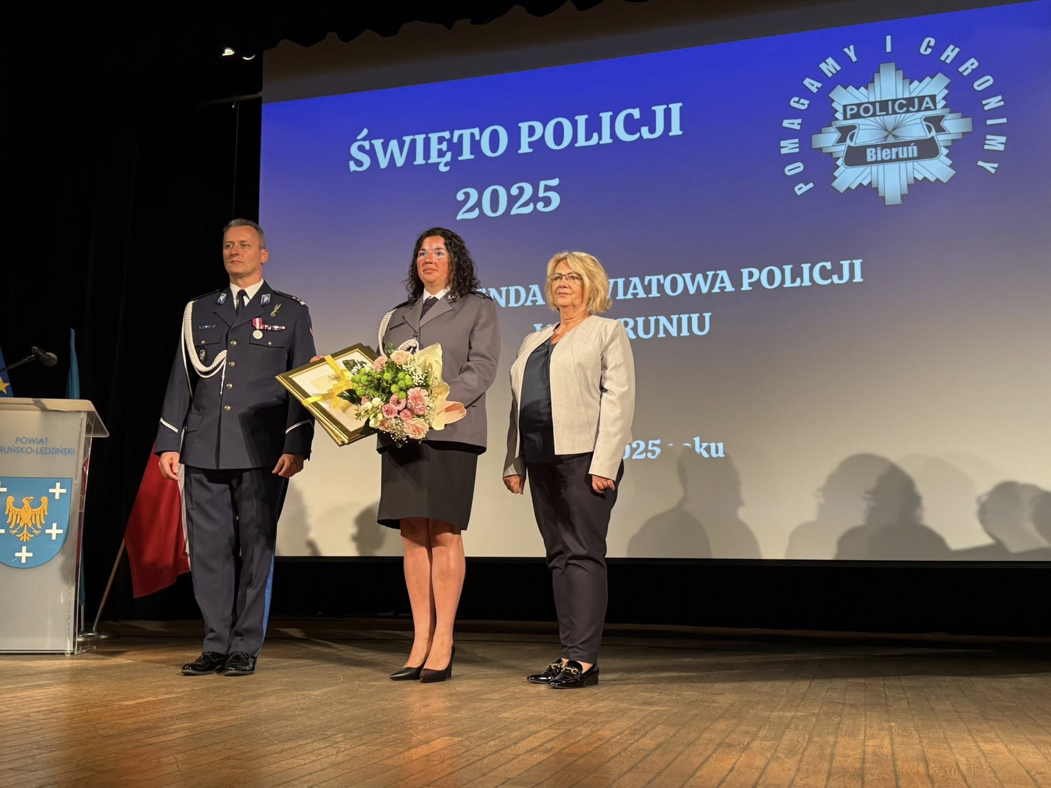 dwie kobiety i mężczyzna stojące na cenie. Jedna z kobiet stoi w mundurze policyjnym a w dłoniach trzyma bukiet kwiatów.