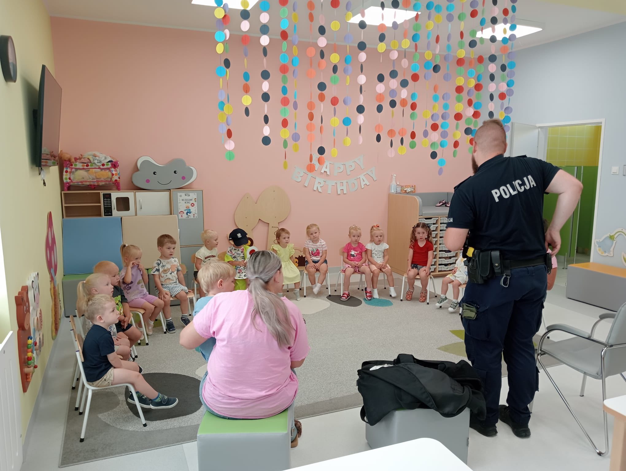 Dzieci siedzące w sali zajęć i wizytujące je policjant