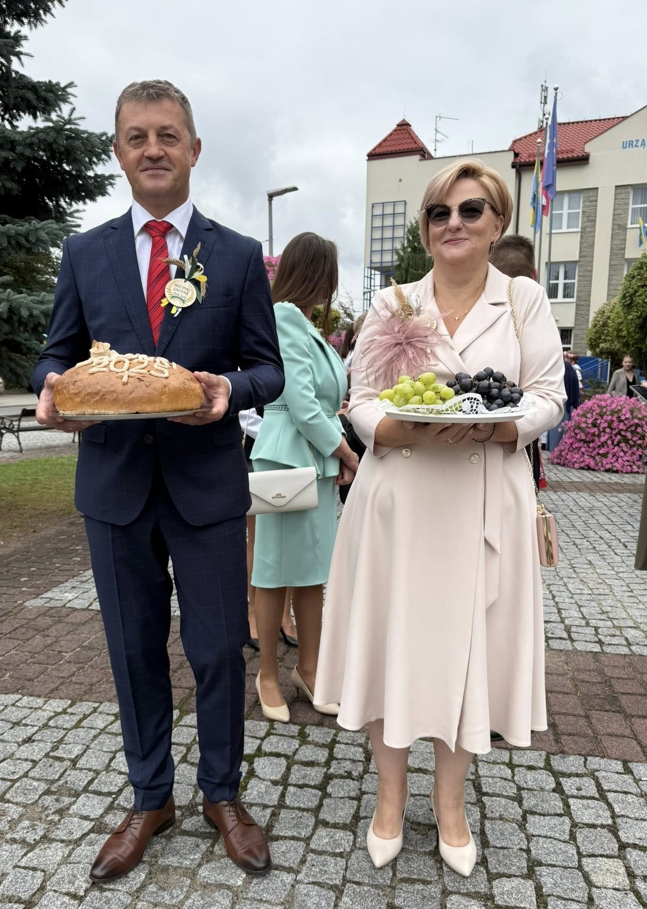 Starości dożynkowi - kobieta i mężczyzna stojący przed budynkiem urzędu z chlebem i winogronami 