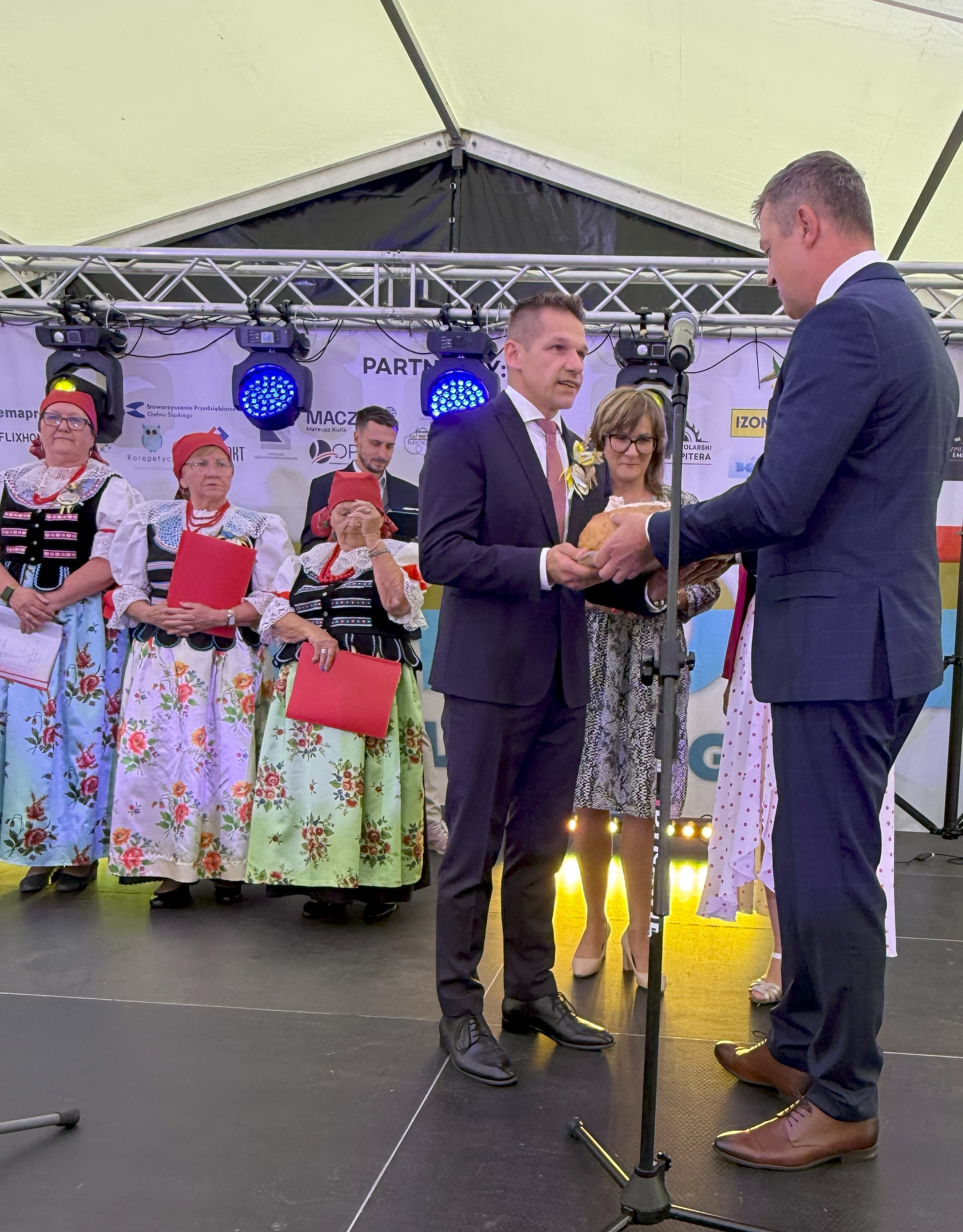 Na scenie kobiety w strojach ludowych i dwaj mężczyźni przekazujący sobie chleb 